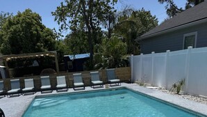 Ein rechteckiger Swimmingpool mit klarem, blauem Wasser, umgeben von einem weißen Zaun und einer überdachten Sitzgruppe mit Stühlen.