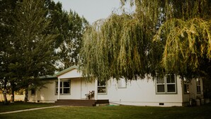 A single-story house with a front porch, a small garden, and a large tree in front. - Cheerful Country Cottage (Rexburg)