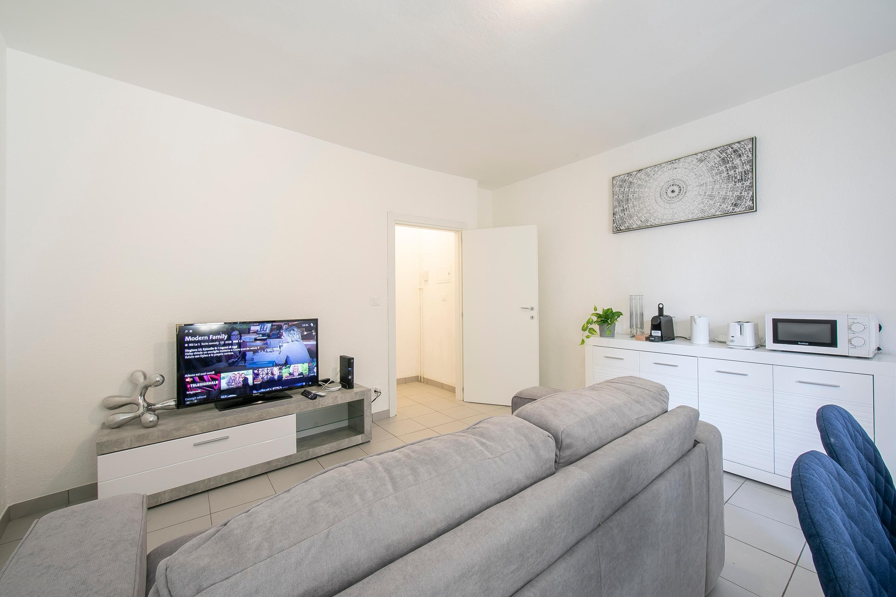 A modern living room with a grey sofa, a flat-screen TV, a coffee machine, and a microwave on a white cabinet.