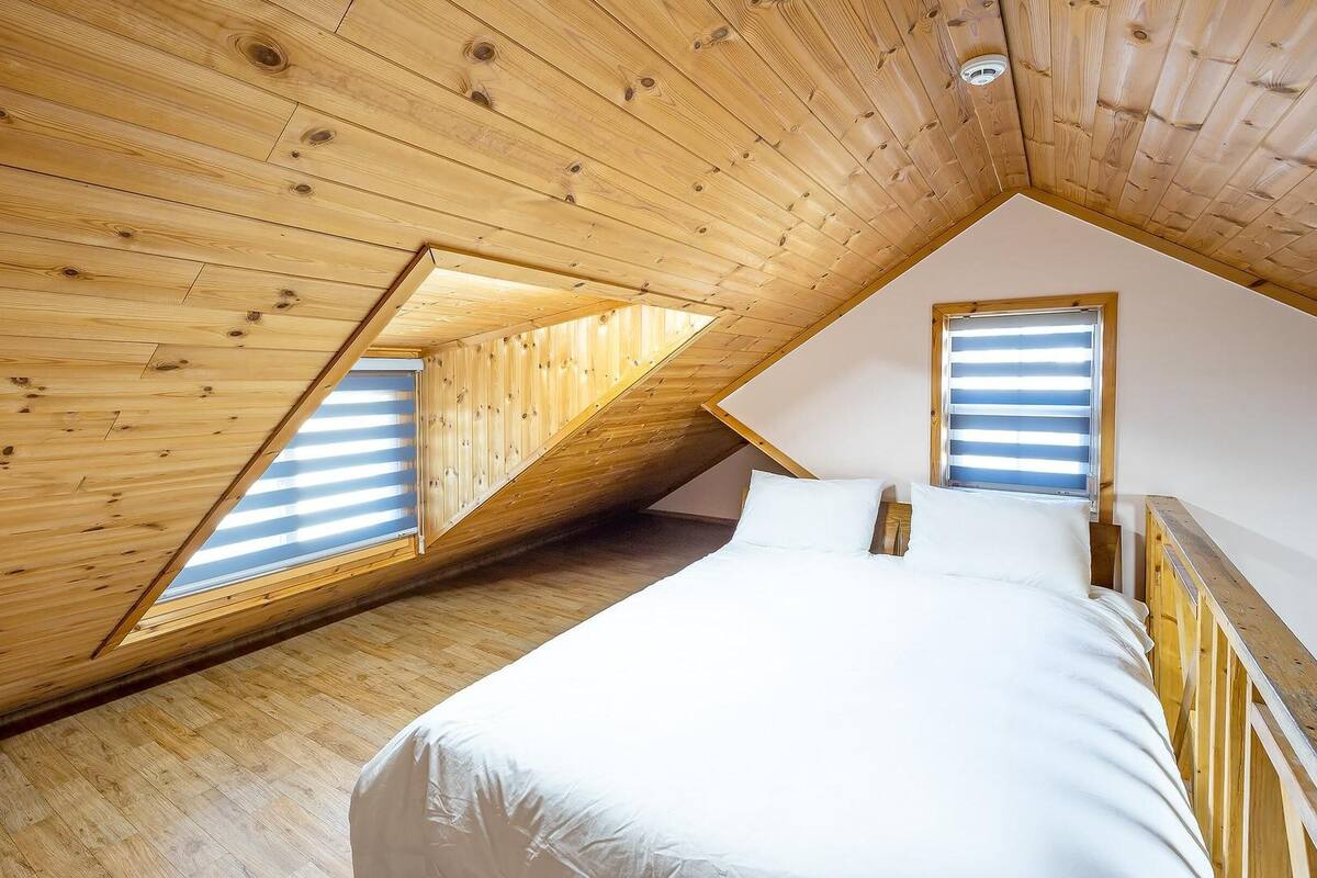 Une chambre avec un plafond en pente, des murs en bois et deux fenêtres équipées de stores.