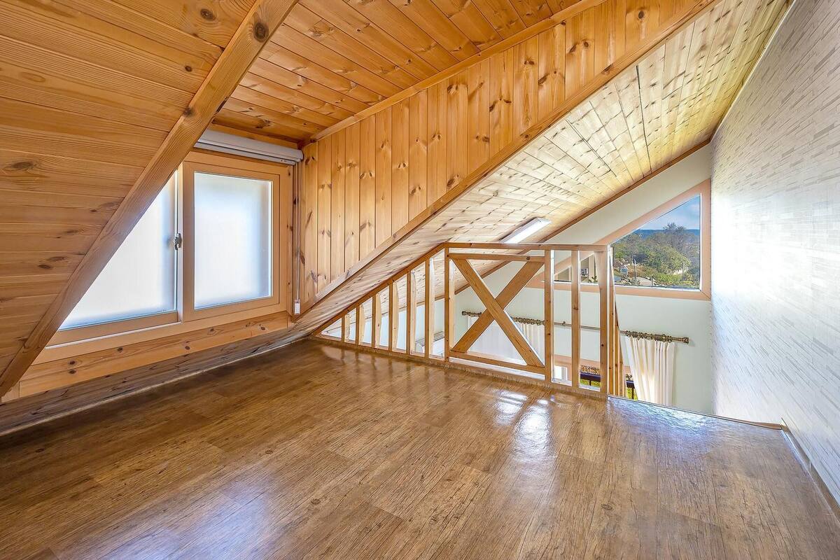 Une pièce mansardée spacieuse, dotée de planchers en bois, d’un plafond en pente et de grandes fenêtres offrant une vue sur l’extérieur.