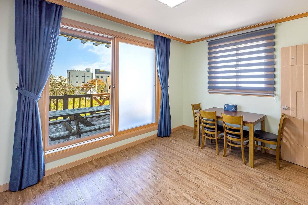 Une pièce avec un plancher en bois, une table entourée de chaises, une fenêtre avec des stores et une porte.