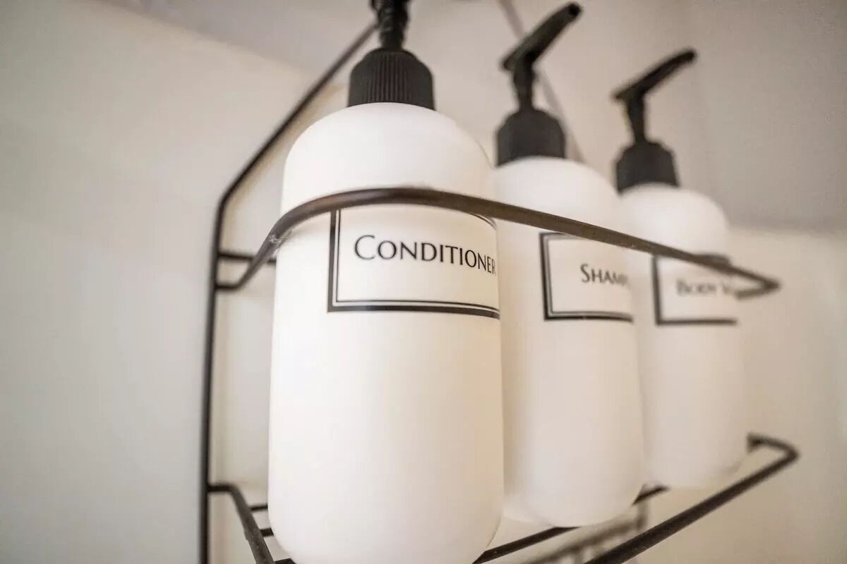 Une étagère en métal sur laquelle sont posées trois bouteilles blanches étiquetées « APRÈS-SHAMPOING », « SHAMPOING » et « CRÈME ».