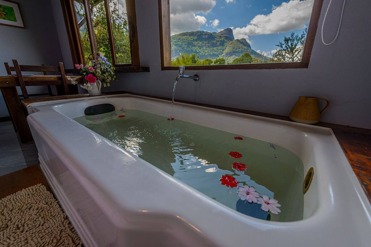 Un baño con una bañera, una ventana con vistas a la montaña, una silla de madera y un florero con flores.
