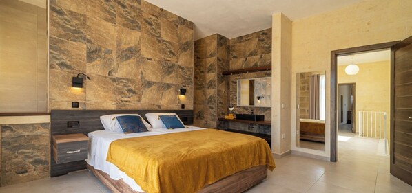 A modern bedroom with a large bed, a nightstand, a dresser, a bathroom with a sink and mirror, and a door leading to another room.