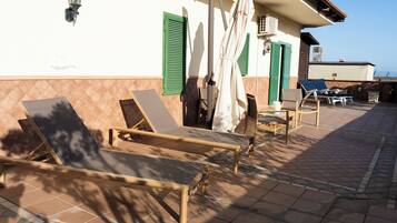 A sunlit outdoor seating area with lounge chairs and a table, a tiled floor, and a building with green shutters and a tiled roof.