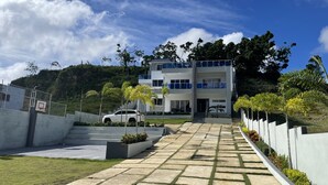Un moderno edificio de varios pisos con un camino pavimentado que conduce a la entrada, rodeado de jardines cuidados y un paisaje montañoso de fondo.