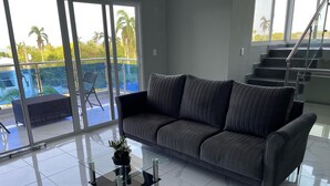 Una sala de estar moderna con un sofá gris, una mesa de centro de cristal y vistas a un balcón con piscina y palmeras.