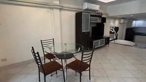Een ruime woonkamer met een ronde glazen tafel en vier stoelen, een plafondventilator en een kitchenette op de achtergrond.