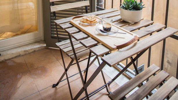 Una pequeña zona de comedor al aire libre con mesa y bancos, una planta en maceta y vistas a través de una ventana con barrotes.