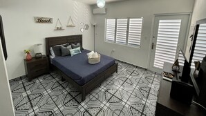 A bedroom with a bed, bedside tables, a lamp, a wall-mounted shelf with decorative items, a window with blinds, and a door.