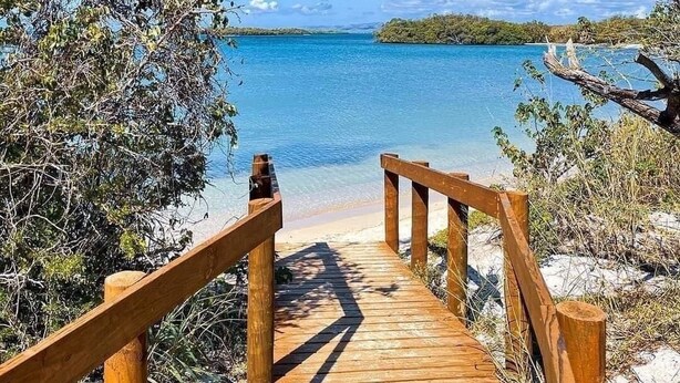 A wooden boardwalk leading to a beach with clear blue water and a distant island.