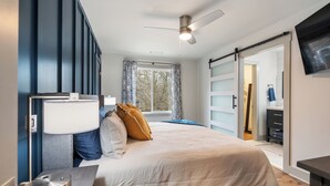 Une chambre à coucher comprenant un lit, une table de chevet, une lampe, un ventilateur de plafond, une fenêtre avec des rideaux et une porte coulissante donnant sur une autre pièce.