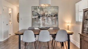 A dining area with a wooden table, four grey chairs, a large painting, and a modern pendant light.
