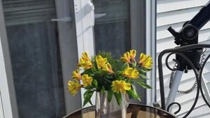 A round glass table with metal chairs on a balcony, a vase with yellow flowers, and a window with blinds. - The Seapointe House. DISCOVER CAPE MAY. (North cape may)