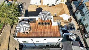 A multi-story building with a rooftop terrace featuring two large black and white striped umbrellas, a wooden deck, and a view of adjacent buildings.