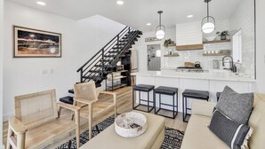 A modern living room with a sofa, chairs, a coffee table, and a staircase leading to a kitchen with white cabinets and a bar area. - Remodeled Rooftop Deck Home | Casino & Ocean Views (Avalon)