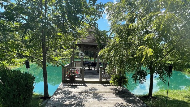 Un belvédère doté d’une terrasse en bois qui mène à un lac, le tout entouré d’arbres et de verdure.