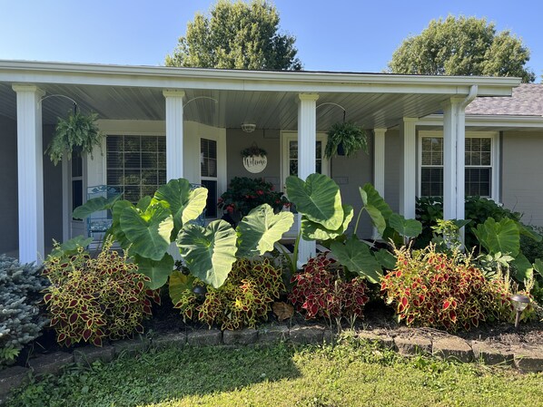 Een huis met een veranda, grote planten en een welkomstbord.