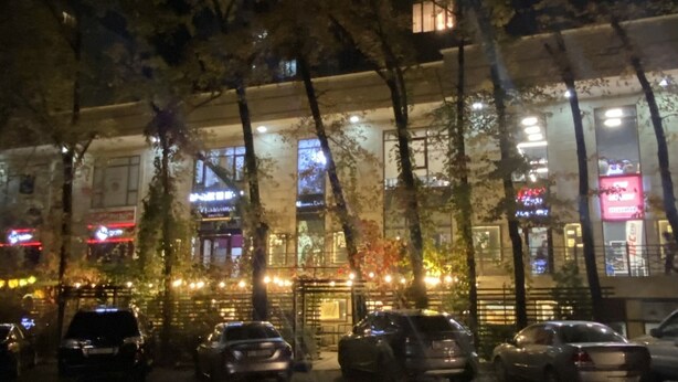Une vue nocturne d’un immeuble avec de nombreuses fenêtres illuminées et un parking où sont garées plusieurs voitures.