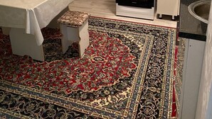 Une cuisine compacte équipée d’une cuisinière à gaz, d’un four et d’un petit réfrigérateur. On y trouve une table recouverte d’une nappe et un banc, ainsi qu’un tapis à motifs sur le sol.