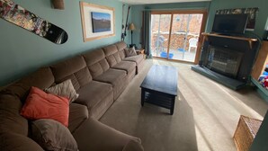 A living room with a sectional sofa, a small coffee table, a fireplace, and a large window with a view of the outdoors. - Condo near wiffletree chairlift! (Carrabassett Valley)