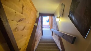 A wooden staircase with a handrail, a wall-mounted light fixture, and a view of the outside. - Condo near wiffletree chairlift! (Carrabassett Valley)