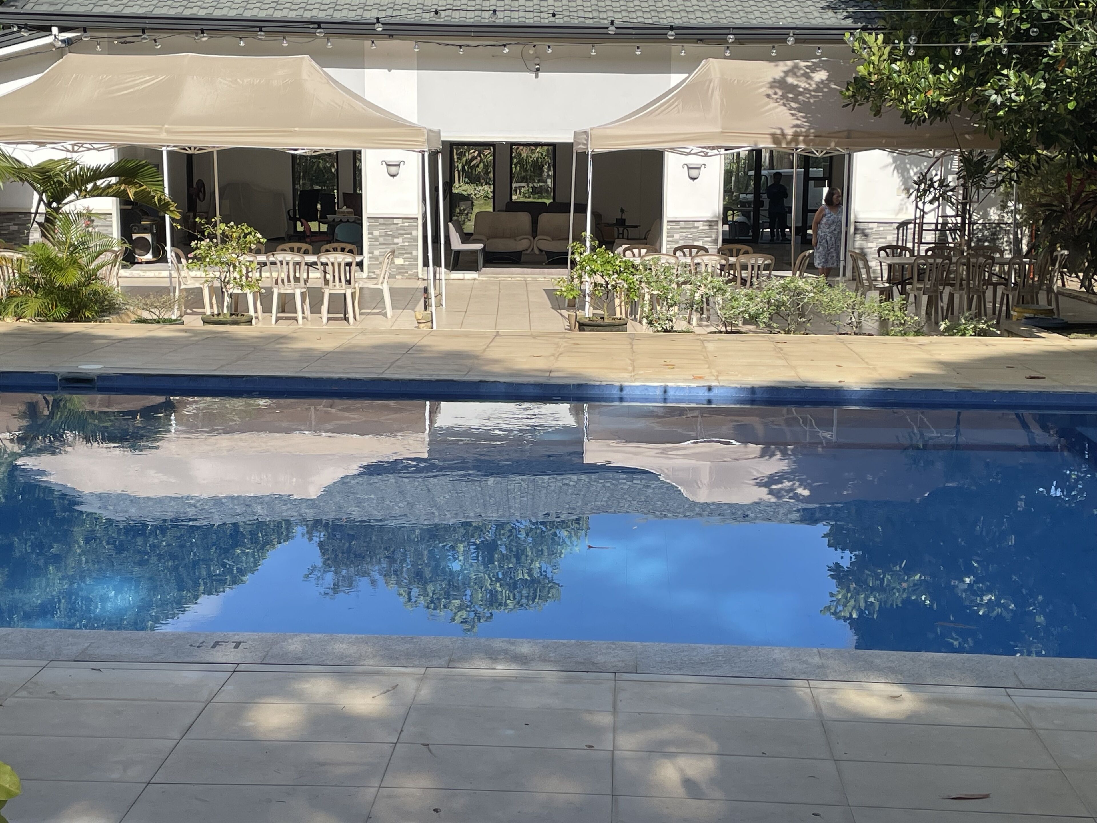 Zona de piscina con solárium con baldosas, mobiliario exterior bajo toldos y vista del edificio al fondo.
