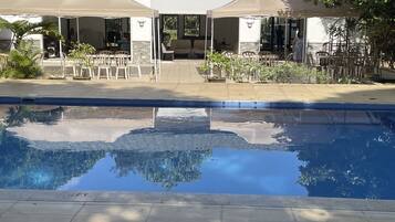 Zona de piscina con solárium con baldosas, mobiliario exterior bajo toldos y vista del edificio al fondo.