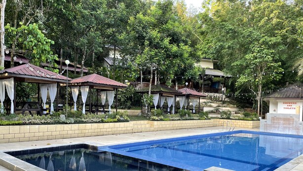 Una piscina rectangular con borde alicatado, rodeada por un muro de ladrillo bajo. Hay zonas de estar cubiertas con cortinas blancas y un cartel que indica duchas y baños.
