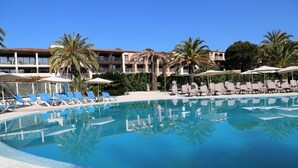 Un espace piscine avec des chaises longues et des parasols, des palmiers et, au fond, un bâtiment doté de balcons.