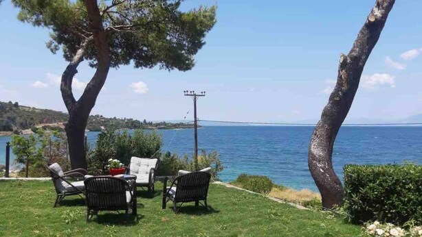 Un espacio al aire libre con césped, dos sillas de mimbre y una mesa, con vista a un cuerpo de agua bajo un cielo despejado.