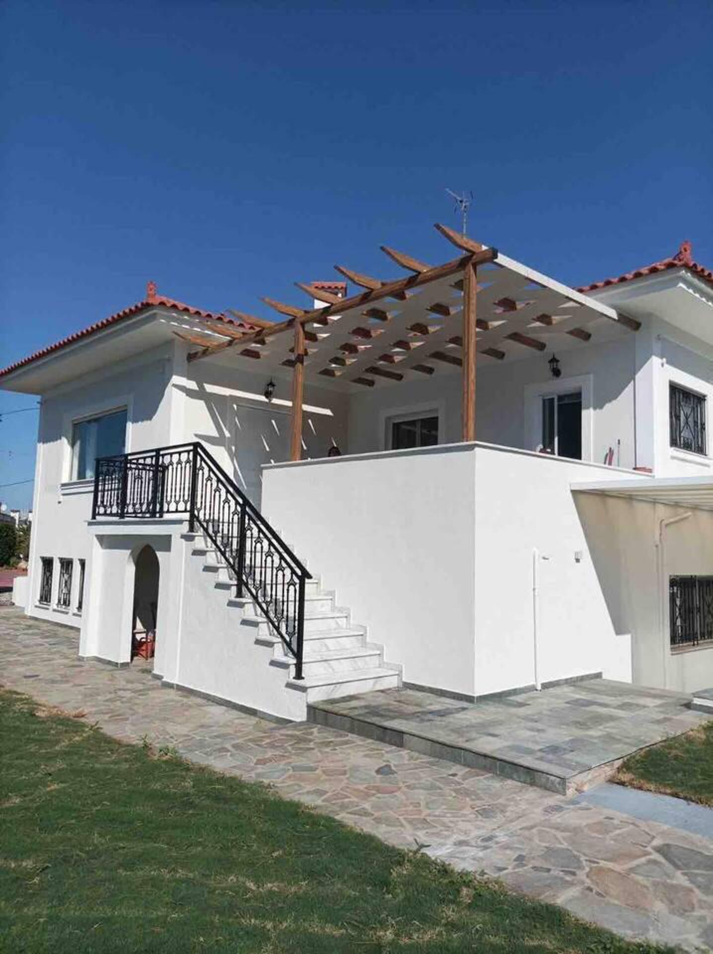 A two-story house with a white exterior, a red-tiled roof, and a wooden pergola on the upper floor. There is a staircase leading to the upper level, and a small garden area in front.