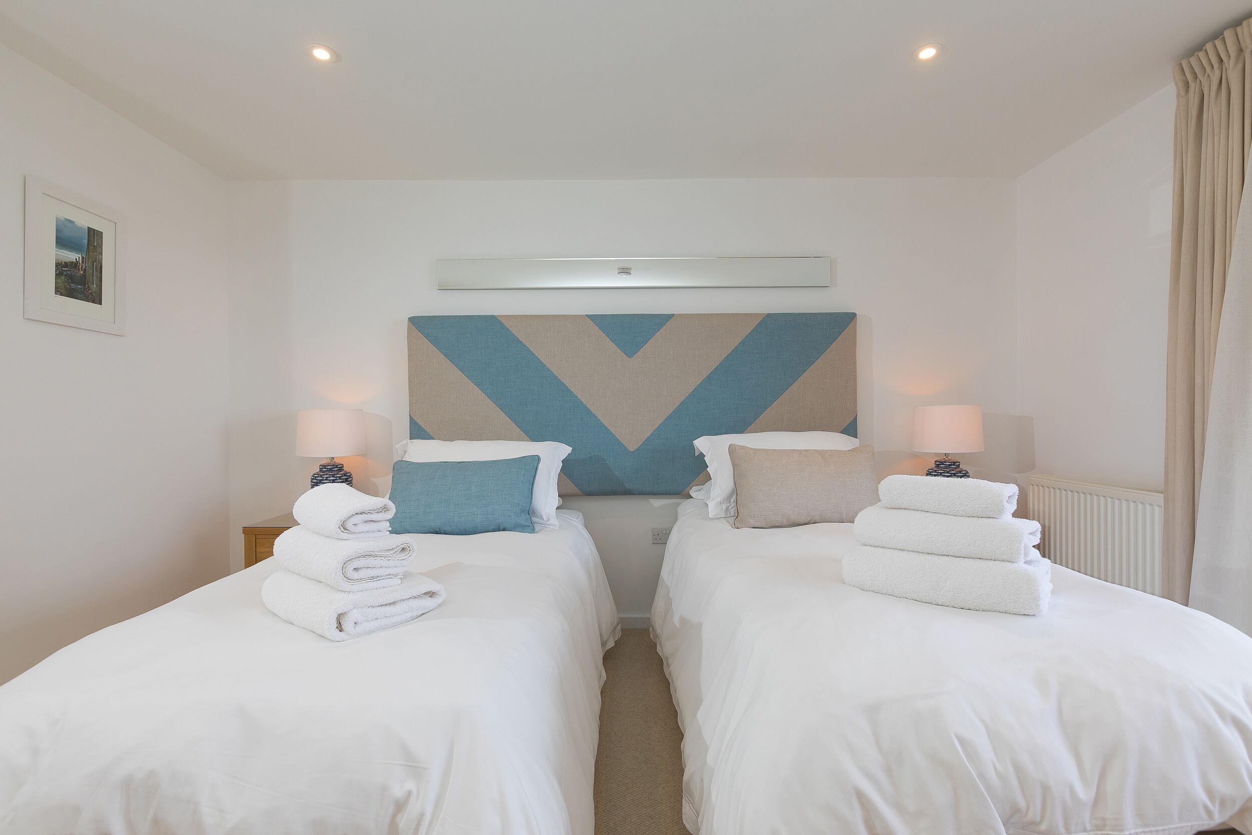 Une chambre d’hôtel avec deux lits, chacun agrémenté d’une serviette pliée, d’une lampe fixée au mur et d’un tableau encadré accroché au mur.