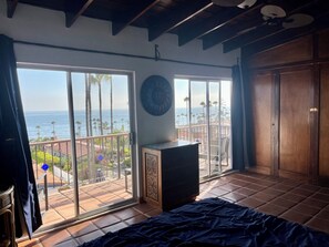 A room with a large sliding glass door leading to a balcony with a view of the ocean and palm trees.