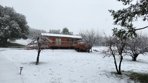 A wooden cabin with a deck, surrounded by snow-covered ground and trees.