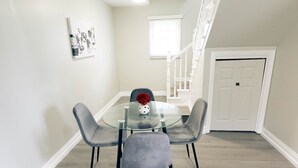 Un comedor con una mesa de vidrio y cuatro sillas grises, un jarrón pequeño con flores rojas, y una escalera que sube a otro nivel.