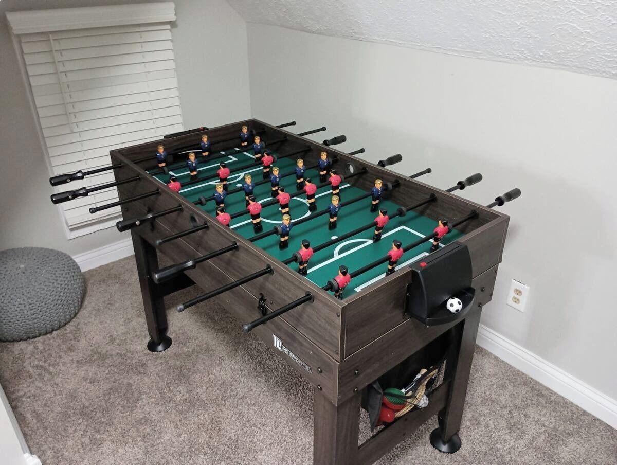 Een tafelvoetbaltafel in een ruimte met vloerbedekking, een raam met jaloezieën en een klein rond tapijt.