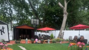 Ein Garten mit Rasenfläche, Kübelpflanzen, einem roten Sonnenschirm und Gartenmöbeln.