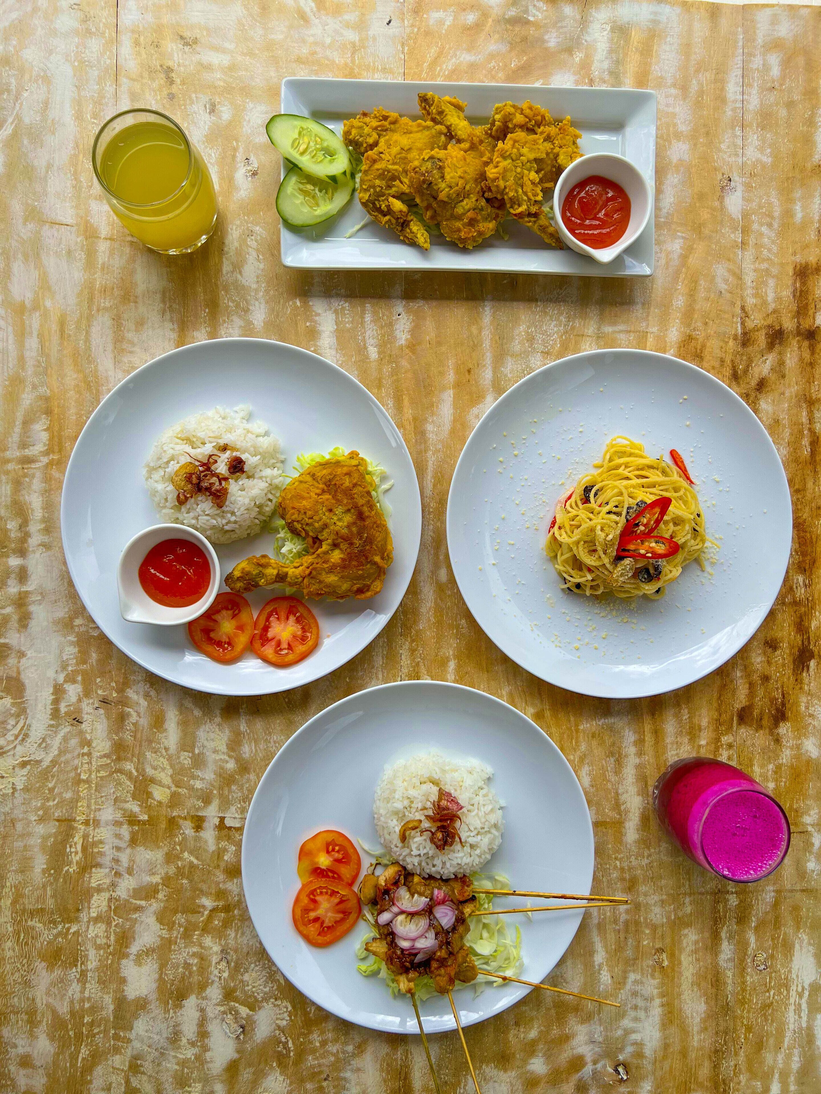 A table with various dishes including fried chicken, rice, spaghetti, and a skewer dish, along with a glass of juice and a glass of pink drink.