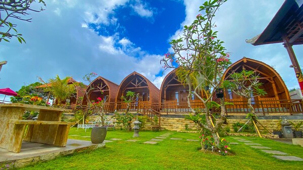 A row of wooden cabins with a stone pathway leading to them, surrounded by lush greenery and a clear blue sky.
