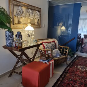 Uma sala de estar acolhedora com uma mesa de centro de madeira, uma poltrona de vime, um banquinho vermelho e um tapete estampado.