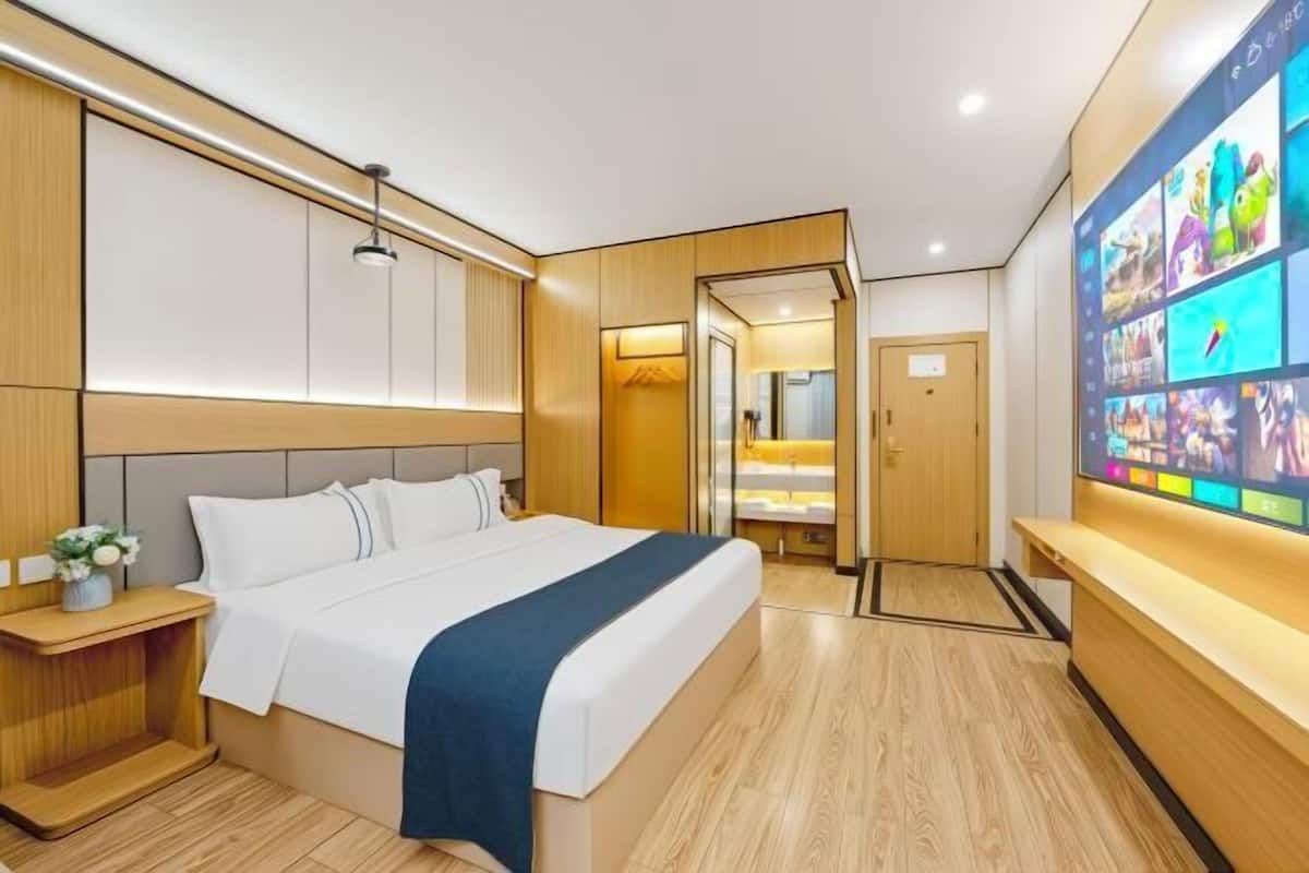 A modern hotel room with a large bed, wooden paneling, a TV wall, and a small desk with a lamp.