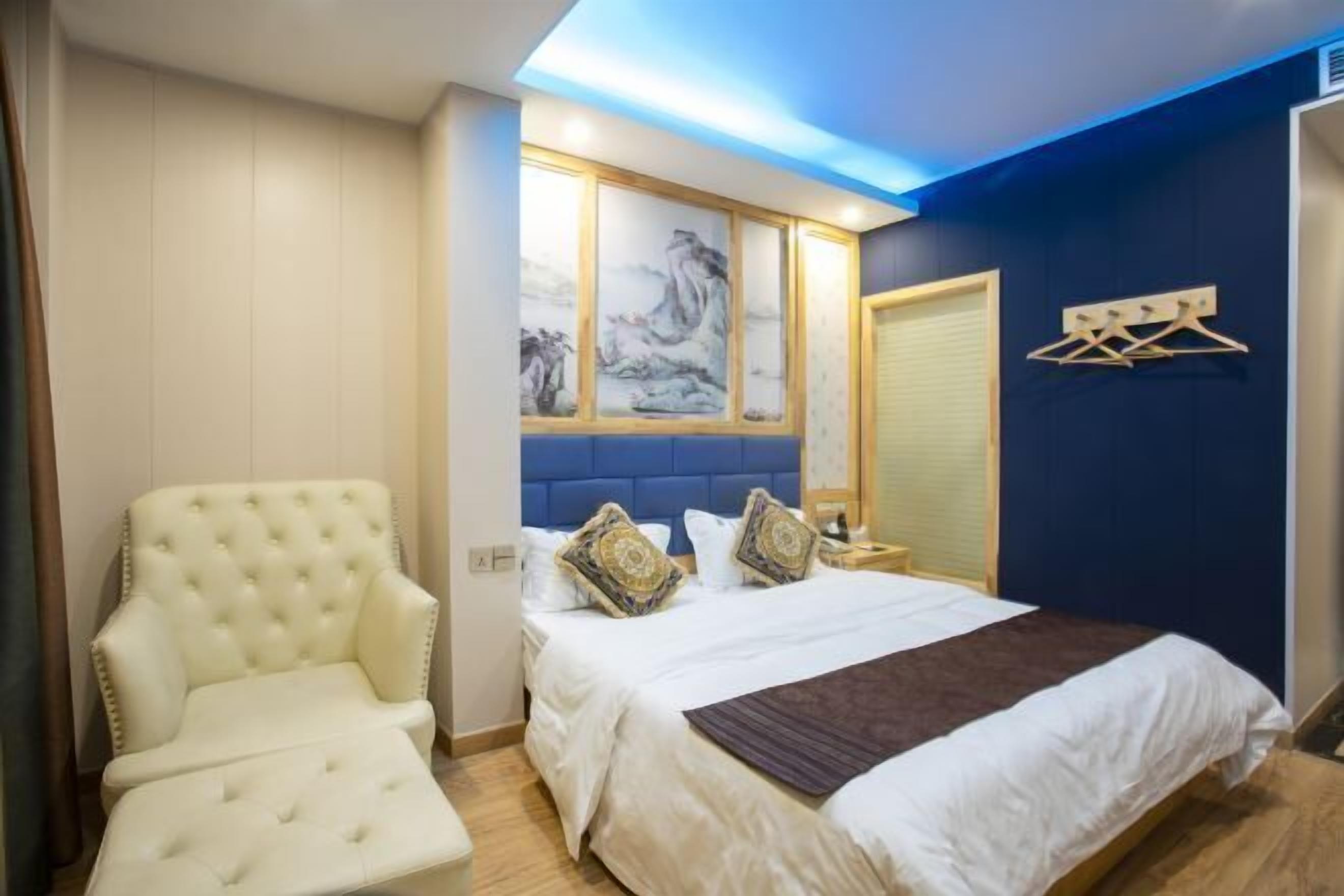A hotel room with a large bed, a white armchair, a blue accent wall, and a painting above the bed.
