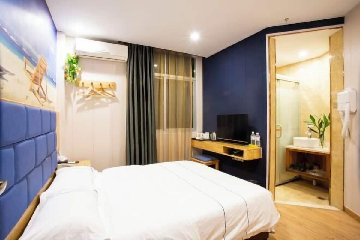 A hotel room with a large bed, a desk with a TV, a window with curtains, and a bathroom visible through an open door.