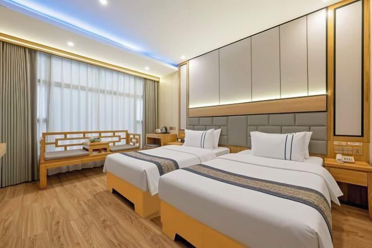 A hotel room with two beds, a wooden bench, a nightstand, and a large window with curtains.
