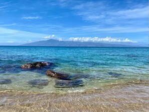 Ein Strand mit Felsen im flachen Wasser, eine entfernte Bergkette und ein klarer Himmel.