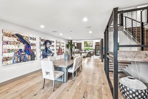 A spacious dining area with a long wooden table and white chairs, a kitchen with modern appliances, and a staircase with a metal railing.