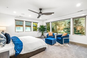 A modern bedroom with a bed, two blue armchairs, a ceiling fan, large windows, and a view of greenery outside.