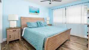 A bedroom with a wooden bed, two bedside tables with lamps, a ceiling fan, a beach-themed painting, and sheer curtains. - ⭐BEACHSIDE BLISS!⭐Singer Island 2/2⭐Huge Pool+Spa (West Palm Beach)